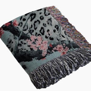 Op de afbeelding: Een opgevouwen sprei met een grijze achtergrond met zwarte luipaardvlekken en roze bloemaccenten. De deken heeft een gefranjerde rand met een mix van kleuren en een donkergrijze franje langs de onderrand.