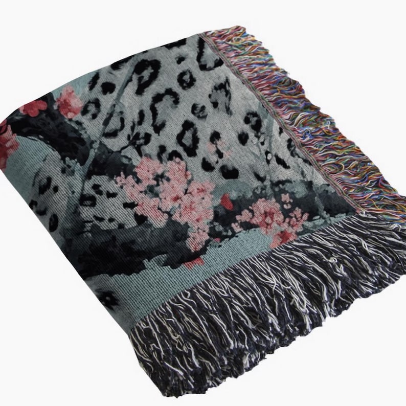 Op de afbeelding: Een opgevouwen sprei met een grijze achtergrond met zwarte luipaardvlekken en roze bloemaccenten. De deken heeft een gefranjerde rand met een mix van kleuren en een donkergrijze franje langs de onderrand.