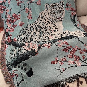 Op de afbeelding: Een geweven deken in teal met een sneeuwluipaardontwerp, rustend op een tak met roze bloesemdetails. De luipaard heeft zwarte vlekken en kijkt de kijker aan. De deken heeft een gefranjerde rand en kan op een bank of bed worden gebruikt.