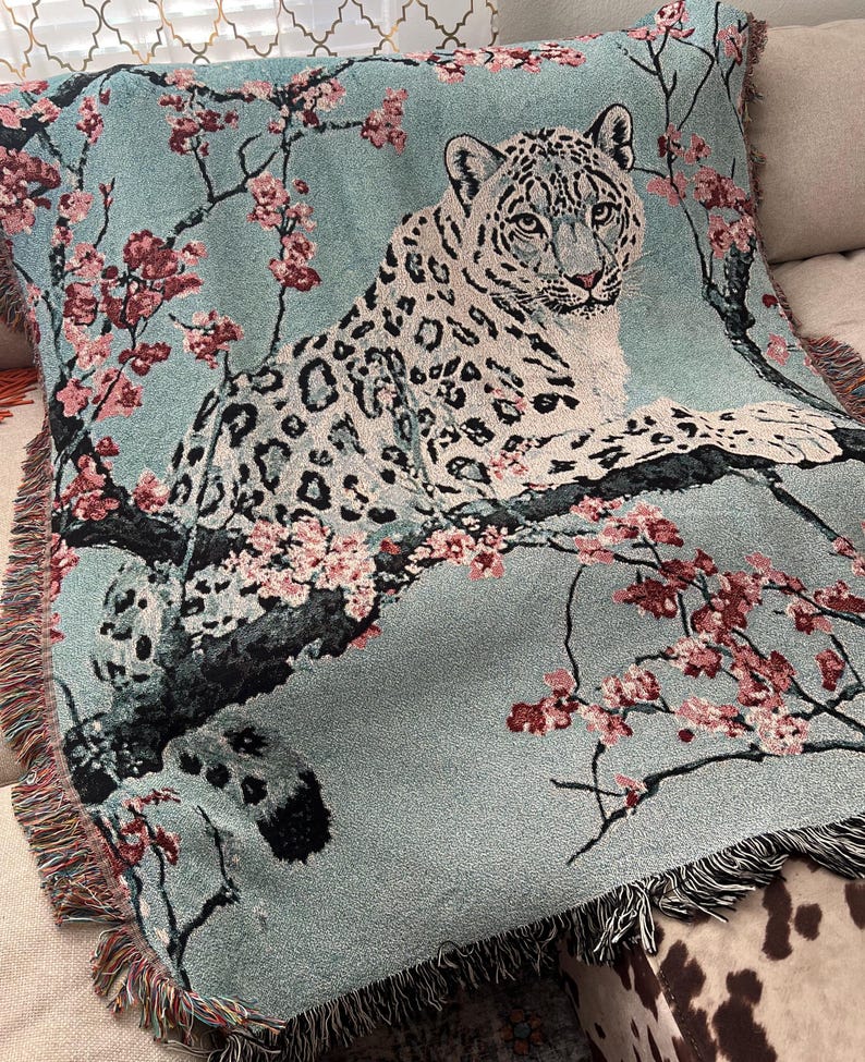 Op de afbeelding: Een geweven deken in teal met een sneeuwluipaardontwerp, rustend op een tak met roze bloesemdetails. De luipaard heeft zwarte vlekken en kijkt de kijker aan. De deken heeft een gefranjerde rand en kan op een bank of bed worden gebruikt.