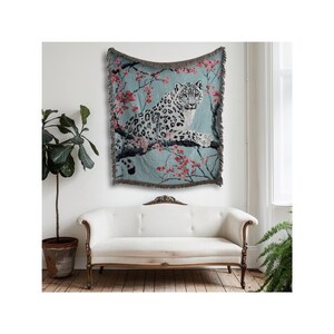 Op de afbeelding: Een geweven sprei met een sneeuwluipaard op een tak met roze bloesems tegen een lichtblauwe achtergrond. De sprei heeft een gefranjerde rand en wordt aan een muur boven een witte bank getoond.