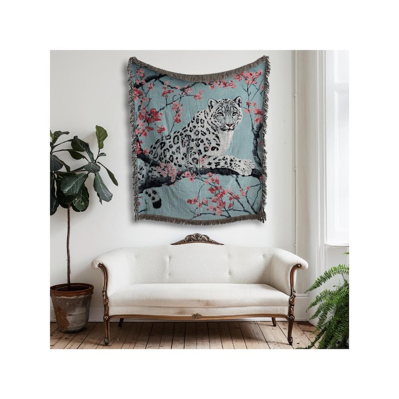 Op de afbeelding: Een geweven sprei met een sneeuwluipaard op een tak met roze bloesems tegen een lichtblauwe achtergrond. De sprei heeft een gefranjerde rand en wordt aan een muur boven een witte bank getoond.