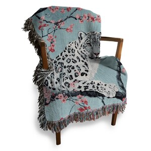 Op de afbeelding: Een lichtblauwe sprei over een houten stoel. De sprei heeft een sneeuwluipaardontwerp met roze kersenbloesems. De sprei heeft een gefranjerde rand. De stoel heeft een houten frame.