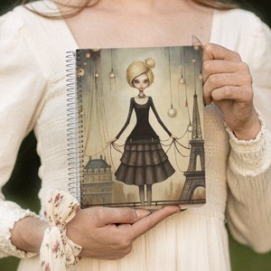 May include: A spiral bound notebook with a colorful illustration of a girl in a black dress standing on a rooftop with the Eiffel Tower in the background. The illustration is in a whimsical style with a dark background and light accents.