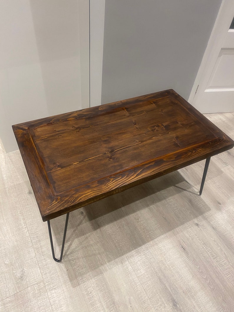 K&ouml;nnte beinhalten: Ein dunkelbrauner Holz-Couchtisch mit einer rechteckigen Tischplatte und einem schlichten Design. Der Tisch hat vier Haarnadelbeine aus schwarzem Metall.