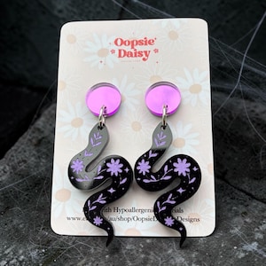 May include: A pair of dangle earrings featuring a black snake-shaped charm with purple floral and star patterns, attached to a circular purple acrylic stud. The earrings are displayed on a beige card with the text Oopsie Daisy printed at the top.