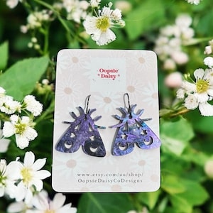 May include: Earrings in the shape of moths, in purple and blue hues, with a starry pattern. The earrings are presented on a card with the text "Oopsie Daisy" and shop details. The background shows white flowers and green leaves.