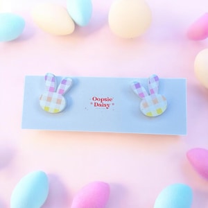 May include: A pair of pastel pink and yellow bunny-shaped earrings with a gingham pattern. The earrings are on a white card with the text "Oopsie Daisy" printed on it.