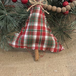 Pu&ograve; includere: Un addobbo natalizio a forma di albero in tessuto imbottito con motivo a quadri rosso, verde e bianco e bordi sfilacciati. L'albero &egrave; sormontato da un piccolo fiocco in corda di juta e ha un bastoncino di cannella marrone come tronco.