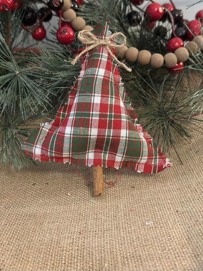 Pu&ograve; includere: Un addobbo natalizio a forma di albero in tessuto imbottito con motivo a quadri rosso, verde e bianco e bordi sfilacciati. L'albero &egrave; sormontato da un piccolo fiocco in corda di juta e ha un bastoncino di cannella marrone come tronco.
