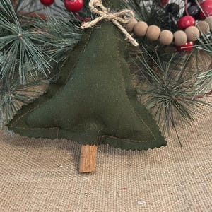 Pu&ograve; includere: Una decorazione per albero di Natale in tessuto verde scuro imbottito, di forma triangolare con bordi sfilacciati. L'albero presenta un piccolo tronco fatto con un bastoncino di cannella marrone alla base e un fiocco in corda di iuta.