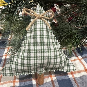 Pu&ograve; includere: Un ornamento in tessuto triangolare a forma di albero, caratterizzato da un motivo a quadri verde e bianco con bordi sfilacciati. Un piccolo fiocco in spago di iuta &egrave; legato sulla punta e un bastoncino di cannella marrone funge da tronco.