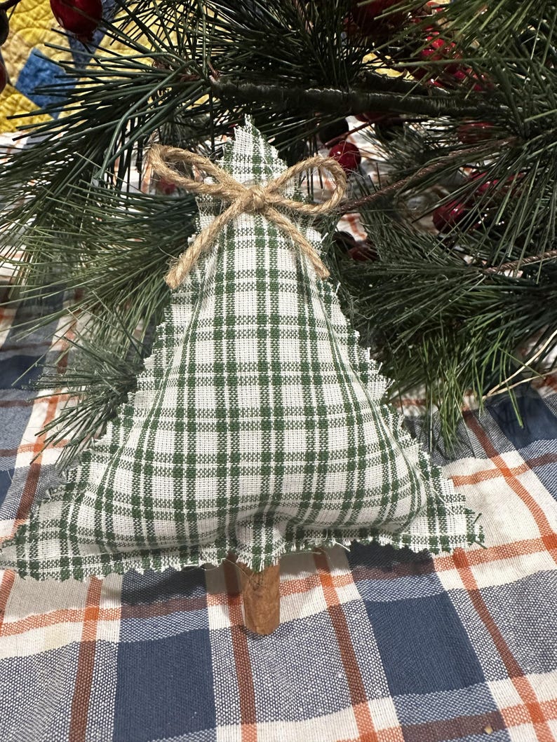Pu&ograve; includere: Un ornamento in tessuto triangolare a forma di albero, caratterizzato da un motivo a quadri verde e bianco con bordi sfilacciati. Un piccolo fiocco in spago di iuta &egrave; legato sulla punta e un bastoncino di cannella marrone funge da tronco.