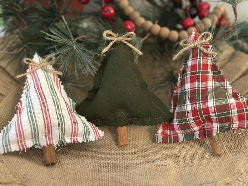 Pu&ograve; includere: Tre decorazioni natalizie a forma di albero in tessuto con bordi sfilacciati e tronchi fatti di bastoncini di cannella. Un albero presenta un motivo a righe bianche e rosse, uno &egrave; in feltro verde scuro e uno ha un design a quadri rossi, verdi e bianchi.