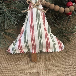 Pu&ograve; includere: Un ornamento per albero di Natale in tessuto imbottito con sfondo bianco e strisce verticali rosse e verdi. I bordi sono sfilacciati e un piccolo fiocco in corda di juta &egrave; legato in cima. Un bastoncino di legno marrone funge da tronco.