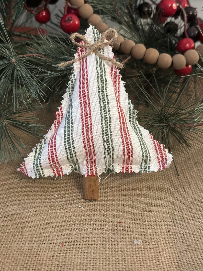 Pu&ograve; includere: Un ornamento per albero di Natale in tessuto imbottito con sfondo bianco e strisce verticali rosse e verdi. I bordi sono sfilacciati e un piccolo fiocco in corda di juta &egrave; legato in cima. Un bastoncino di legno marrone funge da tronco.