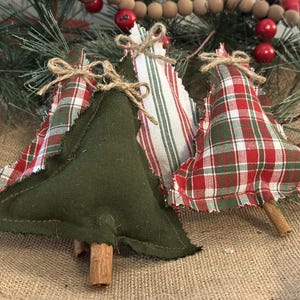 Pu&ograve; includere: Tre decorazioni natalizie a forma di albero in tessuto con un mix di verde oliva tinta unita, motivi a quadri rossi e bianchi e righe. Ogni albero presenta bordi sfrangiati, un piccolo fiocco in spago di juta e un tronco in stecca di cannella.