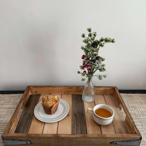 May include: A rustic wooden serving tray with metal corner brackets. A muffin on a white plate and a cup of tea sit on the tray. A small glass vase with greenery and red berries is also on the tray.