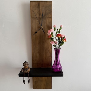 May include: A wooden wall clock with a black shelf. The clock has a dark brown wooden face and black hands. A small figurine sits on the shelf next to a purple vase with pink and red flowers. The clock is mounted on a white wall.
