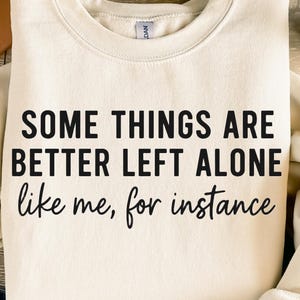 Puede incluir: Sudadera crema con el texto negro "SOME THINGS ARE BETTER LEFT ALONE like me, for instance". Se ve una pequeña planta suculenta en una maceta blanca en el fondo.