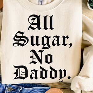 Puede incluir: Sudadera color crema con la frase "All Sugar, No Daddy" impresa en negro con fuente gótica. La sudadera está doblada, con las mangas y el dobladillo inferior visibles. Una pequeña planta suculenta en una maceta blanca está en el fondo.