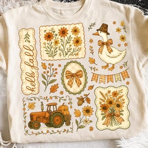 May include: A beige t-shirt featuring a graphic print with the text "hello fall" in cursive. The design includes a white duck wearing a brown hat, an orange tractor with pumpkins, floral arrangements, a bow, and autumn leaves in shades of orange and yellow.