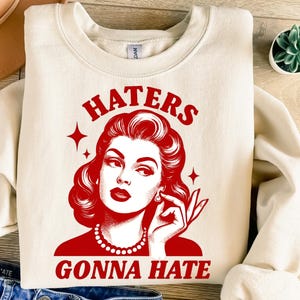 Pode incluir: Camisola creme com um gráfico vermelho de uma mulher e as palavras "HATERS GONNA HATE". O gráfico apresenta uma ilustração em estilo vintage de uma mulher com lábios vermelhos, brincos e um colar de pérolas.