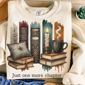 May include: Cream-colored sweatshirt featuring a graphic of books, a candle, and a coffee cup. The design includes the text "Just one more chapter." The books are stacked with a pillow on top. The candle is lit, and steam rises from the coffee cup.