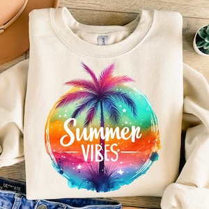 Puede incluir: Sudadera de color crema con un gráfico colorido de una palmera dentro de un círculo con las palabras "Summer Vibes". El gráfico tiene un efecto de acuarela con tonos de rosa, naranja, azul y verde. La sudadera está doblada.