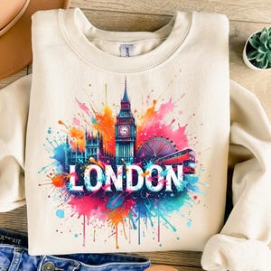 May include: Cream-colored sweatshirt featuring a colorful London design. The design includes Big Ben, the London Eye, and a red double-decker bus, with the word "LONDON" in white. The design has a watercolor splash effect.