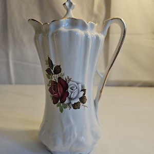 Può includere: Brocca in ceramica bianca con coperchio e manico, con un motivo floreale di rose rosse e bianche. La brocca ha un corpo scanalato e finiture dorate. Un oggetto decorativo per servire bevande.