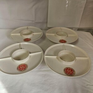 May include: Four off-white plastic serving trays, each featuring a central circular bowl and three divided sections. Each tray includes a circular sticker with the GE logo and text reading Family Day, Open House, Saturday, October 8, 1955.