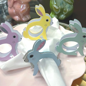 Puede incluir: Cuatro servilleteros de conejito en colores pastel sobre un plato rosa con una servilleta blanca. Los servilleteros son en tonos amarillo, azul, verde y morado. Las orejas de conejo tienen detalles de purpurina. Decoración de mesa de Pascua.