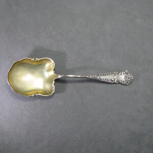 May include: A silver serving spoon with a gold-colored bowl and ornate handle. The spoon has a decorative, floral design on the handle's end. The bowl has a unique, scalloped shape.