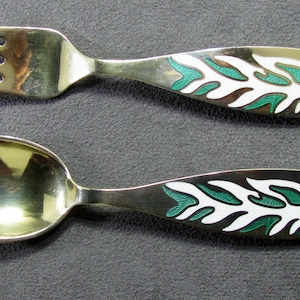May include: A silver fork and spoon set. The handles feature a leaf design in white, green, and blue enamel. The spoon has a rounded bowl, and the fork has four tines. The set is displayed on a gray surface.