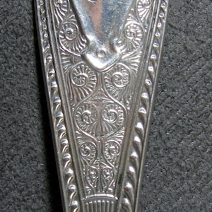 May include: Close-up of a silver spoon with ornate floral and scrollwork detailing. The spoon's bowl features an engraved design with the initials "J H". The handle tapers to a point, showcasing intricate patterns.