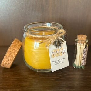 May include: A glass jar candle with a cork lid and a label that reads "PURIFIED BY BEES 100% Organic Beeswax Hand poured in NJ NET WT. (5oz)". The candle is a light yellow color and is sitting on a wooden surface. A small glass jar with matches is next to the candle.
