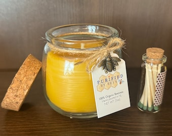 May include: A yellow beeswax candle in a clear glass jar with a cork lid, tied with twine and a bee charm. A white tag reads Purified by Bees, 100% Organic Beeswax, Hand poured in NJ, NET WT. (5oz). A small glass vial of matches sits beside it.