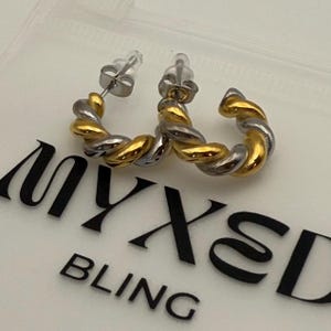 May include: A pair of gold and silver hoop earrings with a twisted design. The earrings are on a white background with the text "MYXED BLING".