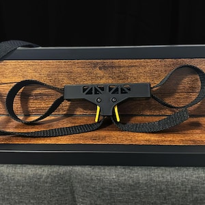 May include: A black and yellow strap system with a geometric design, set against a wood-grain background. The system features a central black component with yellow accents and two loops of black fabric.
