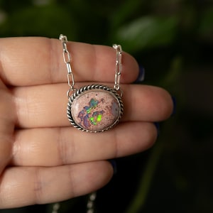 May include: A silver necklace featuring an oval pendant with a pink and multicolored opal stone. The stone has flecks of green, purple, and blue. The pendant is framed by a twisted rope design and hangs from a delicate chain.