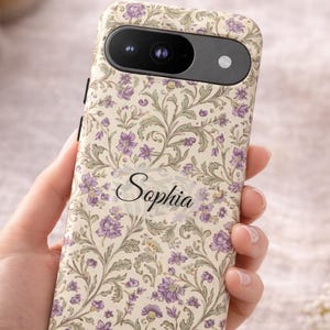 Puede incluir: Funda de teléfono con un estampado floral en crema, verde y morado. La funda tiene el nombre "Sophia" en escritura cursiva. La funda tiene un recorte para la cámara y está diseñada para un smartphone.