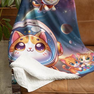 Puede incluir: Una manta de felpa con un diseño temático espacial con gatos de dibujos animados. La manta muestra un gato con un casco de astronauta, un gato alado y otros gatos en un entorno espacial con planetas y estrellas. La parte inferior de la manta es blanca.