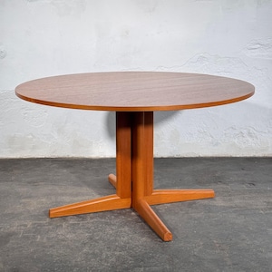 Puede incluir: Una mesa de comedor redonda de madera con un acabado cálido en color marrón medio. La mesa presenta una superficie circular lisa sostenida por una base central que se divide en cuatro patas distintas e inclinadas hacia el suelo.