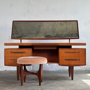 Könnte beinhalten: Mid-Century-Modern-Frisiertisch aus Holz mit rechteckigem Spiegel, zwei Schubladen auf jeder Seite und einer kleinen Mittelschublade. Dazu gehört ein passender runder Hocker mit rosa Samtsitz.