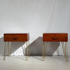 Puede incluir: Dos mesitas de noche de estilo moderno de mediados de siglo. Cada una tiene un mueble rectangular de madera con un cajón y un tirador curvo. Los muebles están sostenidos por cuatro patas doradas de horquilla. La madera es de color cálido.