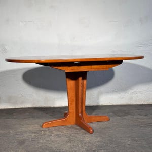 Può includere: Un tavolo da pranzo in legno color marrone medio con un piano di forma ovale e una base a piedistallo centrale dotata di quattro gambe divaricate. La superficie del tavolo è liscia e lucida, mostrando una venatura naturale del legno.