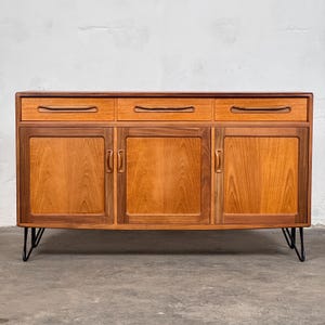 Könnte beinhalten: Ein Sideboard aus Holz im Mid-Century-Modern-Stil mit drei Schubladen und drei Schranktüren. Die Schubladen haben eingelassene Griffe, und die Schranktüren haben Metallgriffe. Das Sideboard wird von schwarzen Metall-Haarnadelbeinen getragen.