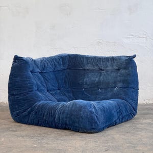 May include: A low-profile, corner-shaped modular sofa piece upholstered in a deep blue velvet fabric. The design features a tufted backrest and seat with visible button detailing, creating a soft, quilted texture across the entire surface.