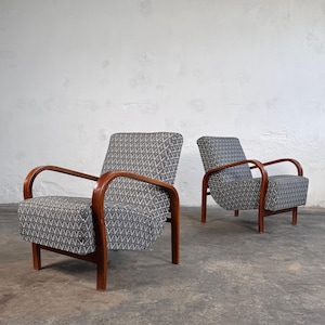 Op de afbeelding: Twee vintage fauteuils met een geometrisch patroon in zwart en wit. De stoelen hebben gebogen houten armleuningen en poten, die een modern design uit het midden van de eeuw laten zien. De achtergrond is een effen witte muur en een grijze vloer.